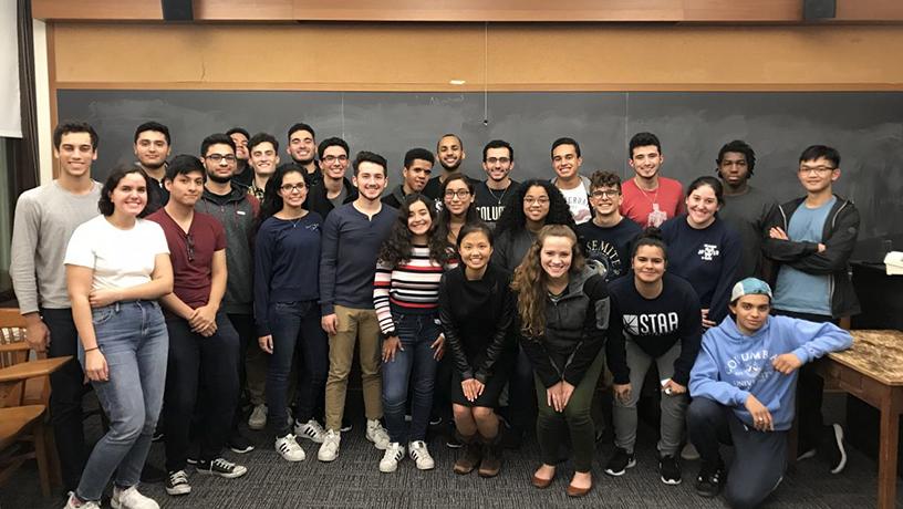 A group of SHPE members stands in front of a blackboard for a group photo.