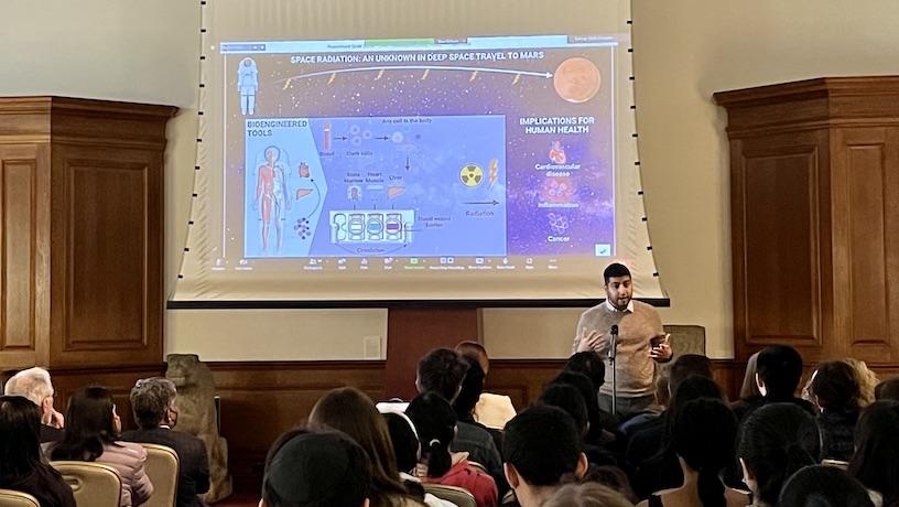 Naveed Tavakol presenting to an audience. The screen behind him reads "Space Radiation: An Unknown in Deep Space Travel to Mars"