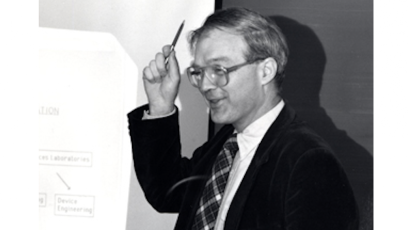 A middle aged white man standing at a black board teaching. He is wearing a suit coat and tie. He is pointing at the screen with a pencil.