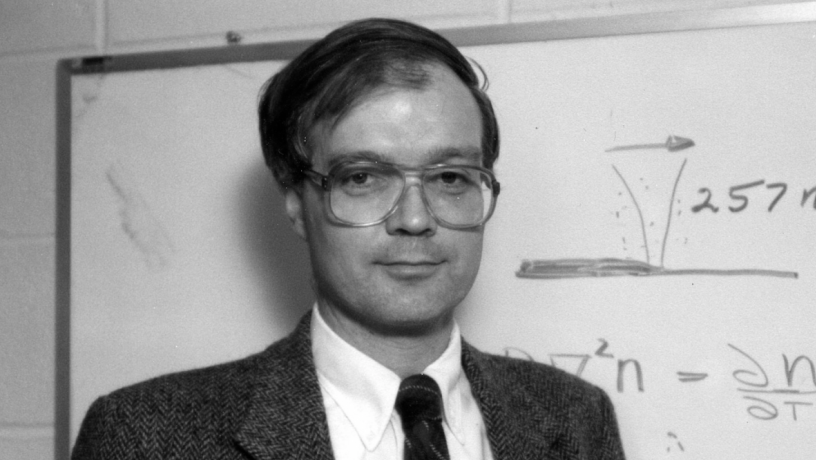 A middle aged white man standing in front of a black board. He is wearing a suit coat and tie. 