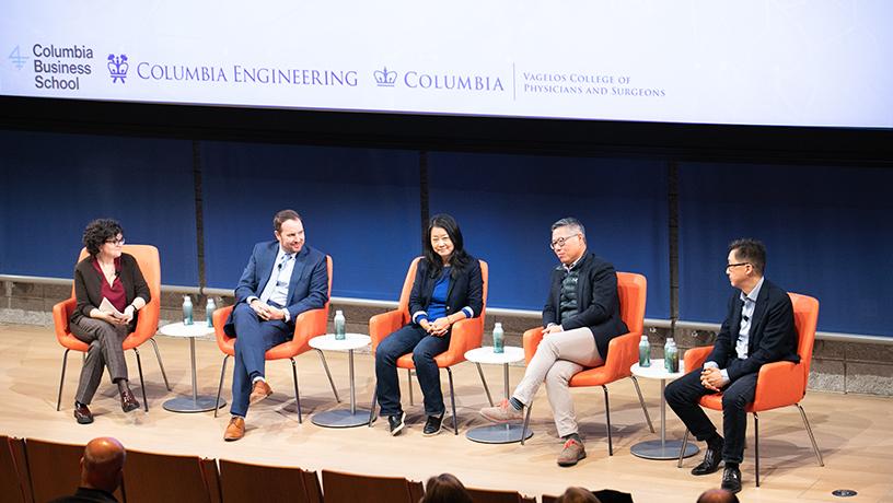 Five panelists, two women and three men, sitting in chairs on stage. Audience members watch in the foreground.
