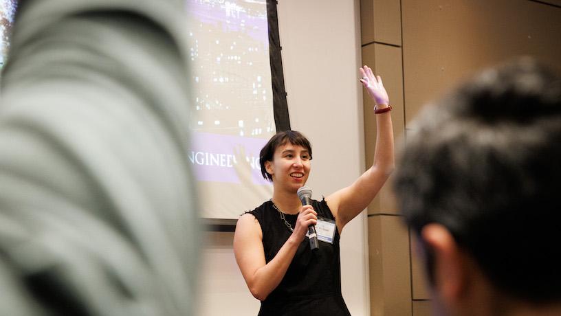 A fast pitch competition contestant holding into a micorophone and raising their hand as they present to an audience. The backs of audience members obstruct a view of the contestant.