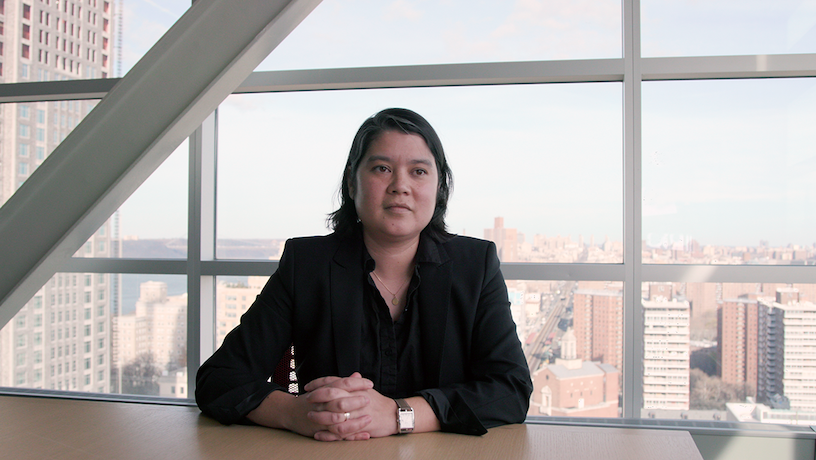 Krystin Myers sitting at a table with hands together. A view of New York City comes through the background window.