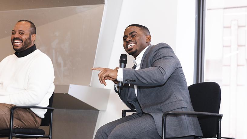 A man sits in a chair on-stage smiling and holding a microphone