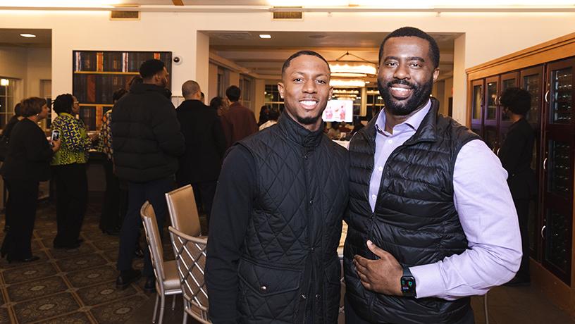 Two people smiling for a photo. Others crowd the room behind them.