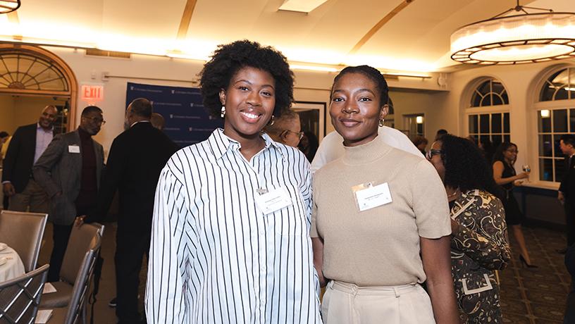 Two people wearing name tags smiling for a photo. Others crowd the room behind them.