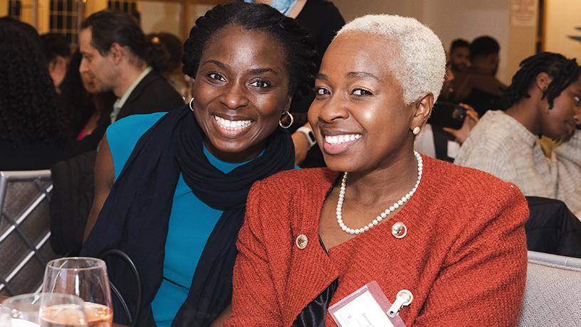 Two people smiling for a photo. Behind them, other people sit at tables.