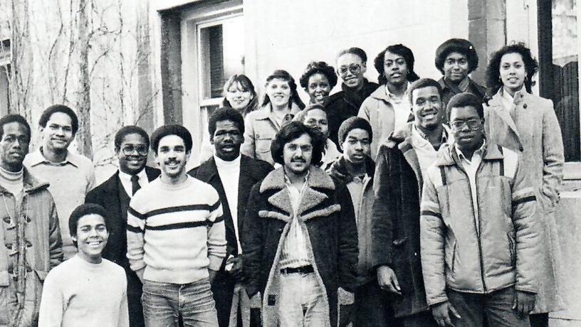 A black-and-white photo of 18 smiling people staggered in rows