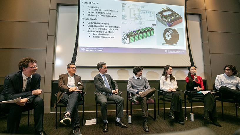 A panel of seven people sitting in chairs beneath a projected screen. The second right-most person speaks into a microphone as the other panelists listen to them.