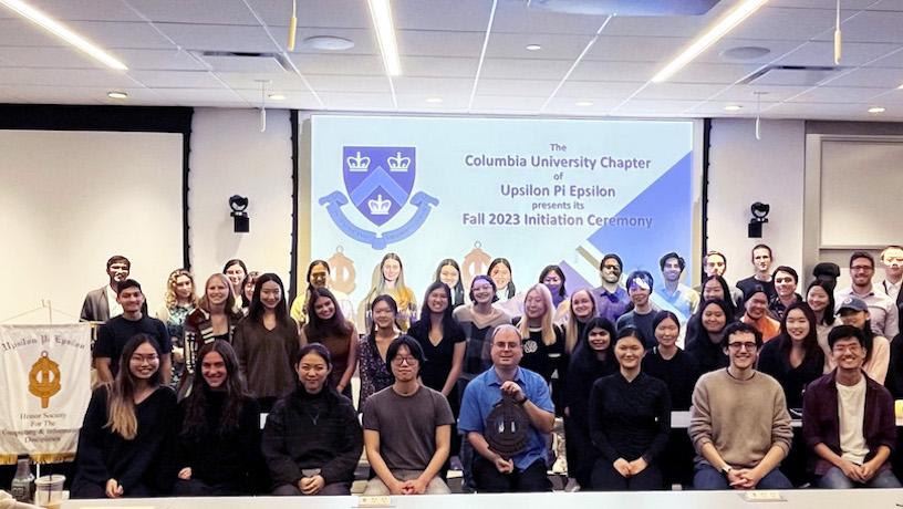 Three rows of people smiling for a photo. In the background, a large screen reads "Columbia University Chapter of Upsilon Pi Epsilon Fall 2023 Initiation Ceremony"